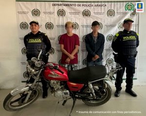 En la imagen se observa los dos capturados junto a dos uniformados de la Policía Nacional. Frente a ellos la motocicleta hurtada. En la parte posterior se observa el banner que identifica al Departamento de Policía Arauca.