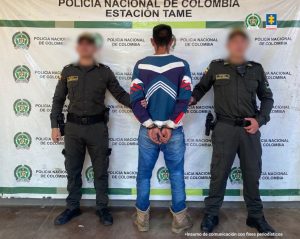 Se visualiza al capturado de espaldas, junto a dos uniformados de la Policía Nacional. Detrás el banner que identifica a la Policía Nacional en Tame-Arauca