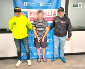 En la imagen se aprecia al capturado con el rostro difuminado junto integrante de la Sijin y un investigador del CTI. Detrás de ellos el banner que identifica a la Fiscalía General de la Nación.