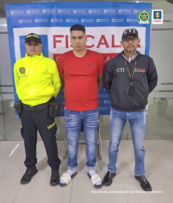 Se observa al capturado junto a un uniformado de la Policía Nacional y un investigador del CTI. En la parte posterior se ubica el banner que identifica a la Fiscalía General de la Nación