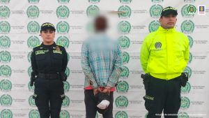 Se visualiza al capturado de espaldas junto a dos uniformados de la Policía. En la parte posterior se observa el banner con los logos que identifican a la Policía Nacional