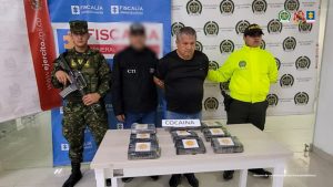  Se visualiza al capturado junto a un investigador del CTI y uniformados del Ejército y Policía Nacional. Frente a la mesa la sustancia alucinógena equivalente 9 kilos de 235 gramos de cocaína. Detrás de ellos los banners institucionales de la Fiscalía General de la Nación, Policía y Ejército Nacional. 