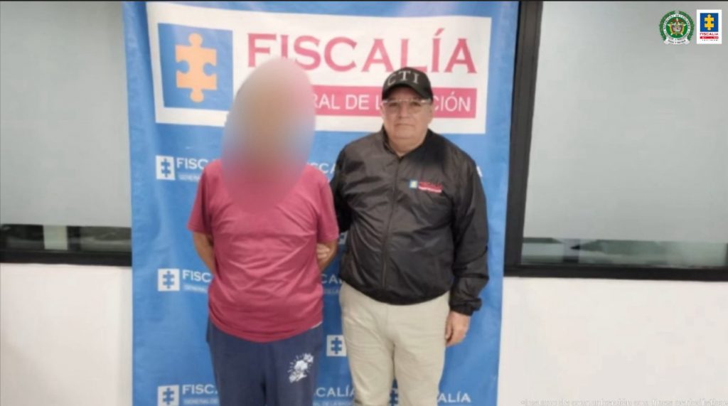 Se visualiza al capturado con el rostro difuminado junto a un investigador del CTI. Detrás se ubica el banner que identifica a la Fiscalía General de la Nación