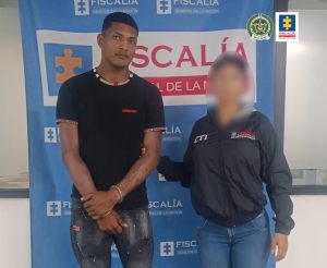 Se visualiza al procesado junto a una investigadora del CTI. Detrás el banner que identifica a la Fiscalía General de la Nación.