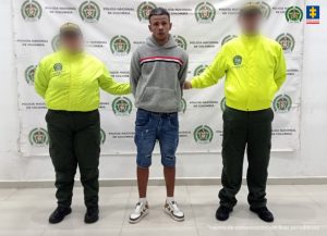  Se visualiza al capturado junto a dos uniformados de la Policía Nacional. Detrás de ellos el banner que identifica a la Policía Nacional.