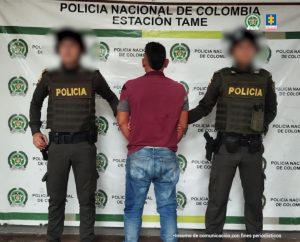 Se visualiza al capturado de espaldas junto a dos uniformados de la Policía Nacional, en la parte posterior se observa el banner que identifica a la Policía Nacional en Tame -Arauca.