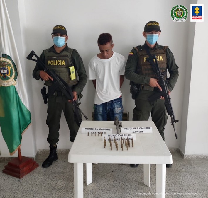 En la fotografía aparece el condenado junto a dos uniformados de la Policía Nacional, a un lado se observa una bandera de la Policía Nacional. Frente a ellos una mesa con munición incautada al momento de la captura