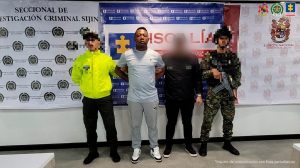 Se visualiza al capturado junto a un investigador del CTI y dos uniformados de la Policía y Ejército Nacional. Detrás de ellos los banners que identifican a la Fiscalía General de la Nación, Policía y Ejército Nacional.