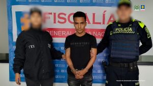 Se visualiza al capturado junto a una investigadora del CTI y uniformado de la Policía Nacional. Detrás el banner que identifica a la Fiscalía General de la Nación.