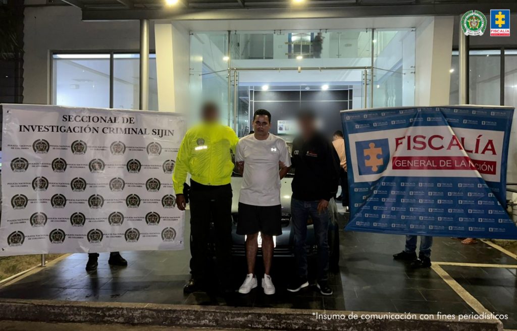 Se visualiza al capturado junto un uniformado de la Policía Nacional y un investigador del CTI. Detrás los banners que identifican a la Seccional de Investigación Criminal Sijín y a la Fiscalía General de la Nación.
