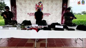 Se visualiza al capturado de espaldas junto a dos uniformados del Ejército Nacional. Frente a ellos una mesa con los explosivos y elementos de intendencia incautados. Detrás el banner que identifica al Ejército Nacional.