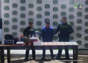 Se visualiza al capturado junto a dos uniformados de la Policía Nacional, detrás se ubica el banner que identifica a la Policía Nacional.