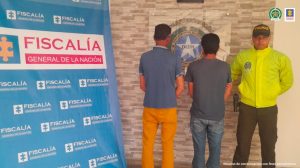 En la fotografía se ve a los dos capturados de espaldas junto a un uniformado de la Policía Nacional. Detrás de ellos se visualizan los banners de la Fiscalía General de la Nación y la Policía Nacional.