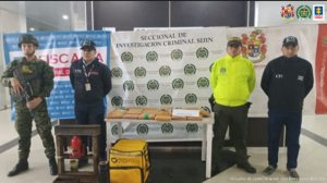Se observa a dos investigadores del CTI y uniformados del Ejército y Policía Nacional, frente a ellos una mesa donde su ubicaron los más de 12 kilos de marihuana incautados. Detrás los banners institucionales de la Fiscalía General de la Nación, Policía y Ejército Nacional.