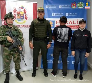 Se visualiza al capturado de espaldas junto a uniformados del Ejército y la Policía Nacional, y una investigadora del CTI. Detrás se ubican los banners institucionales de la Fiscalía, Ejército y Policía Nacional.