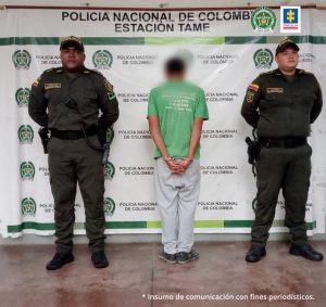 En la imagen aparece el capturado de espaldas junto a dos uniformados de la Policía Nacional. En la parte posterior se observa el banner que identifica a Policía Nacional de Tame (Arauca)