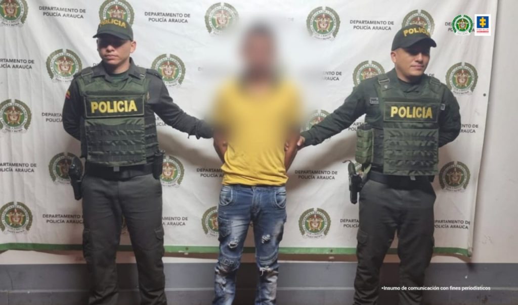 Se visualiza al capturado con el rostro difuminado, junto a dos uniformados de la Policía Nacional. En la parte posterior el banner que identifica a la Policía Nacional.