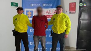 Se visualiza al capturado con el rostro difuminado junto a dos uniformados de la Policía Nacional. Detrás el banner que identifica a la Fiscalía General de la Nación.