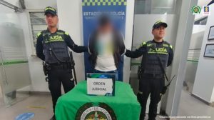 Se observa al capturado con el rostro difuminado junto a dos uniformados de la Policía Nacional. Detrás de ellos el banner que identifica a una estación de Policía en Bogotá.