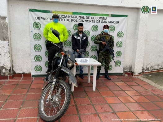 En la fotografía se aprecia al capturado en medio de uniformado de la Policía y Ejército Nacional. Al frente una mesa con elementos materiales y una motocicleta incautada. En la parte posterior un banner del Departamento de Policía de Norte de Santander