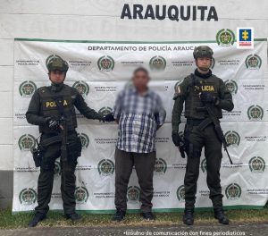 Se observa al detenido con el rostro difuminado junto a dos uniformados de la Policía Nacional. Detrás de ellos el banner que identifica a la Policía en Arauca.