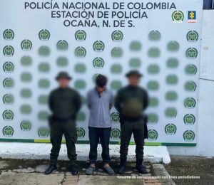 Se visualiza al capturado junto a dos uniformados de la Policía Nacional, detrás se ubica el banner que identifica a la Policía Nacional.