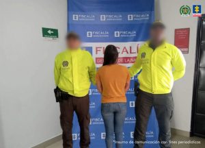 Se visualiza a la capturada junto a dos uniformados de la Policía Nacional con el rostro difuminado. Detrás se ubica el banner que identifica a la Fiscalía General de la Nación.