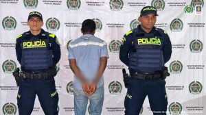 Se observa al capturado con el rostro difuminado junto a dos uniformados de la Policía Nacional. Detrás de ellos el banner que identifica a una estación de Policía en Bogotá.