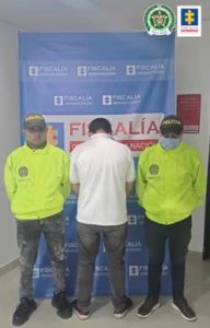 Se visualiza la imagen se visualiza al capturado junto a dos uniformados de la Policía Nacional. En la parte posterior se observa el banner que identifica a la Fiscalía General de la Nación.
