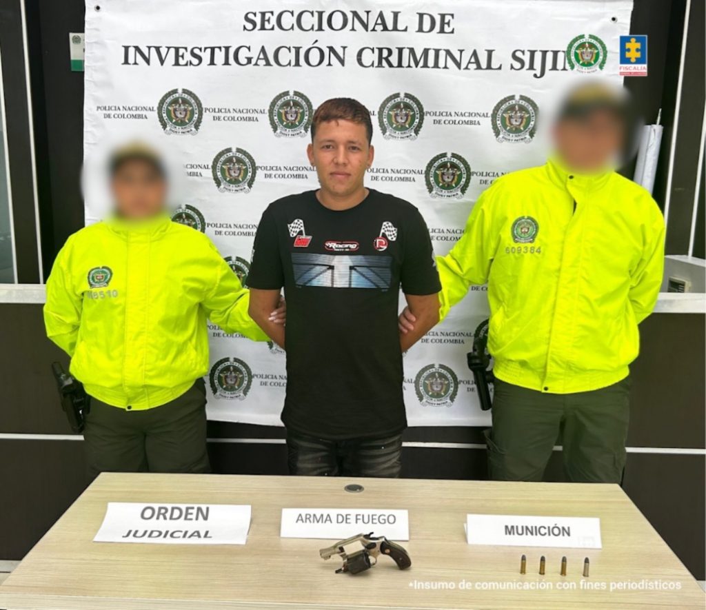 Se observa al capturado junto a dos uniformados de la Policía Nacional. Frente a ellos una mesa con un arma incautada. Detrás de ellos el banner que identifica a la Seccional de Investigación Criminal Sijín de la Policía Nacional.