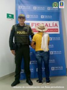 En la imagen se aprecia al capturado de espaladas junto a un uniformado de la Policía Nacional. En la parte posterior se aprecia el banner que identifica a la Fiscalía General de la Nación.
