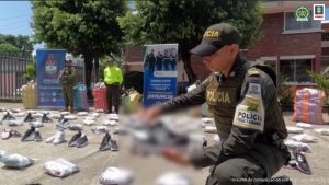 En la imagen se aprecia a un uniformado de la Policía Fiscal y Aduanera mostrando uno de los tenis de imitación. Detrás se observa varios pares de tenis y banner que identifica a la Policía Nacional