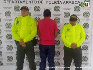 En la imagen aparece el capturado de espaldas junto a dos uniformados de la Policía Nacional. En la parte posterior se aprecia el banner que identifica al departamento de Policía en Arauca