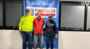 Se visualiza al capturado junto a un uniformado de la Sijin y una investigadora del CTI. Detrás de ellos el banner que identifica a la Fiscalía General de la Nación