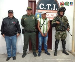 en la imagen se ve una persona detenida bajo cutodia de un investigador del CTI y de la Policía Nacional. detrás suyo un backing de la Fiscalía.