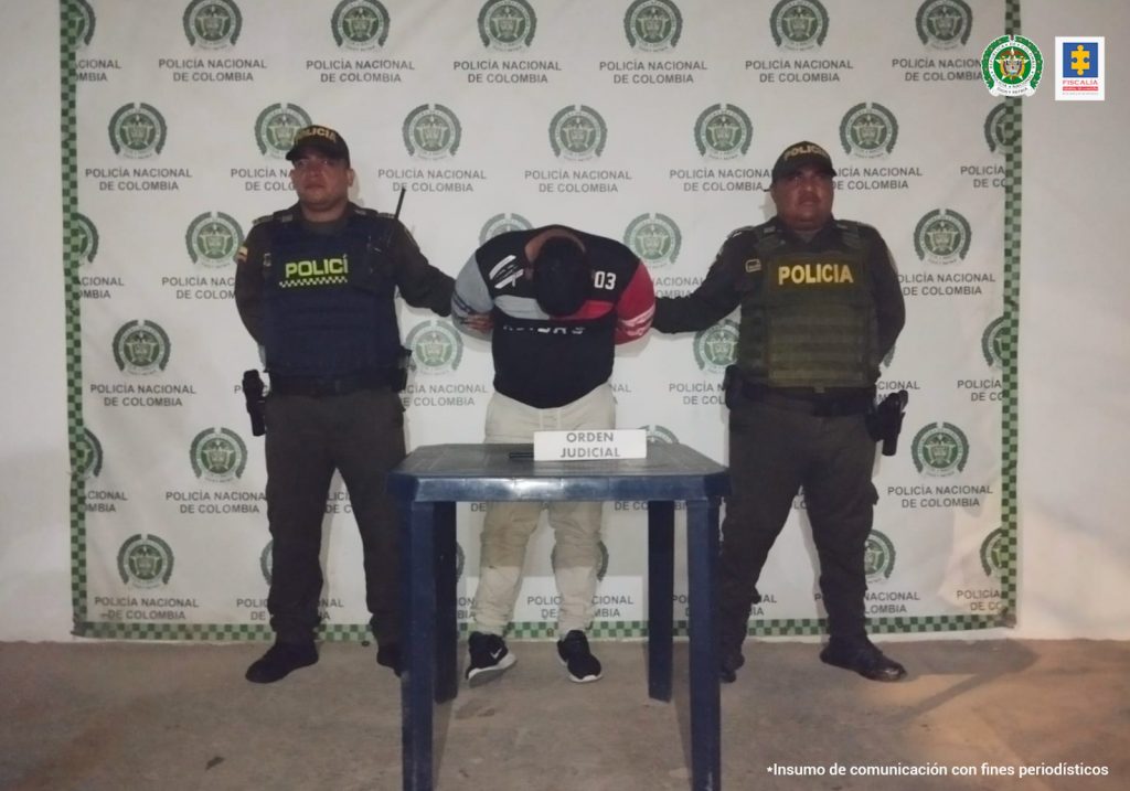 En la imagen se ve una persona detenida bajo custodia de dos uniformados de a Policía Nacional. Detrás suyo un backing institucional.