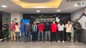 en la imagen se ve un grupo de personas detenidas bajo custodia de la Policía Nacional. Detrás suyo un paredón con el nombre de la Policía del Huila.