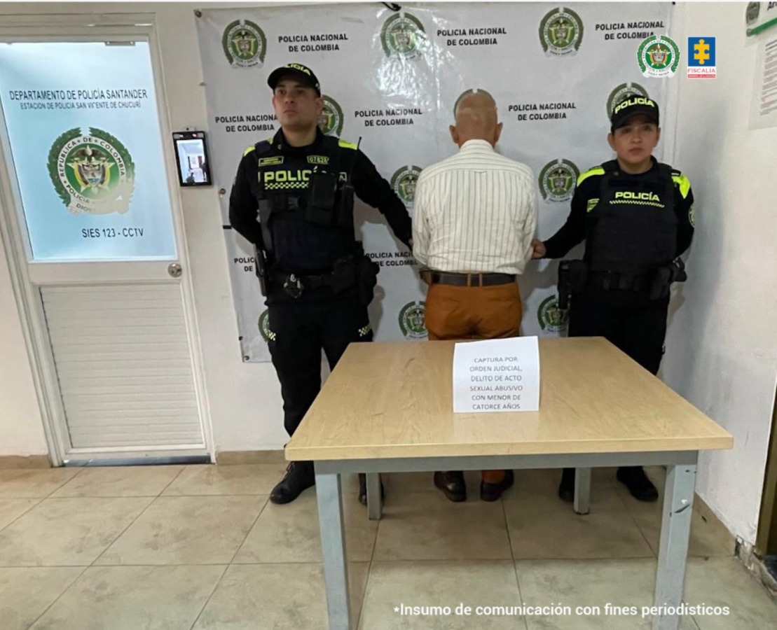Se visualiza al procesado de espaldas junto a dos uniformados de la Policía Nacional. Detrás de ellos el banner que identifica a la Policía Nacional.
