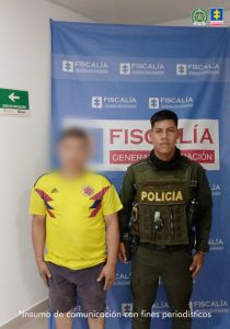 Se observa al capturado con el rostro difuminado junto a un uniformado de la Policía Nacional. Detrás de ellos el banner que identifica a la Fiscalía General de la Nación.