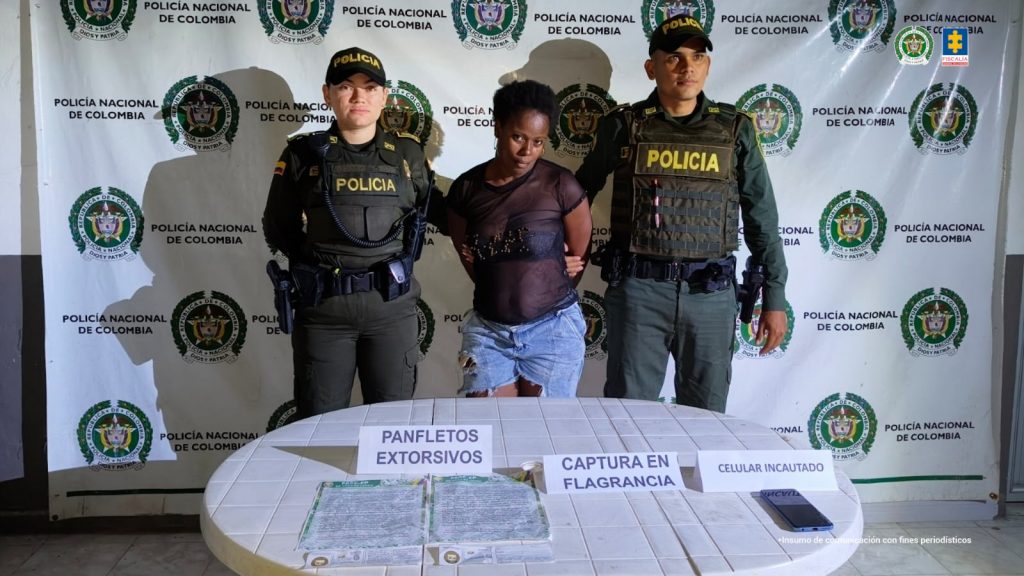 capturada de frente custodiada por dos policías. Detrás banner de la Policía Nacional. Delante de ellos una mesa con material incautado.
