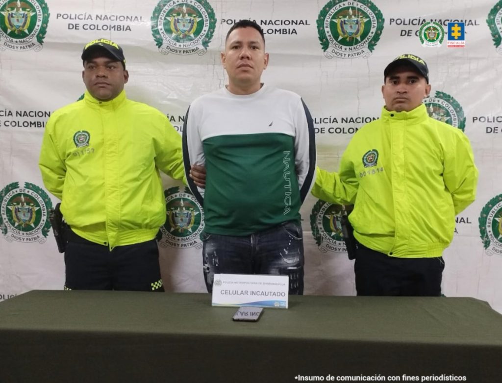 en la imagen se ve una persona detenida bajo custodia de dos uniformados de la Policía Nacional. Delante suyo una mesa con un celular incautado. Detrás, un backing institucional.