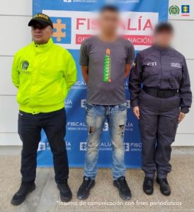 En la imagen se ve un hombre detenido bajo custodia de un investigador del CTI y un uniformado de la Policía Nacional. Detrás suyo, un backing institucional.