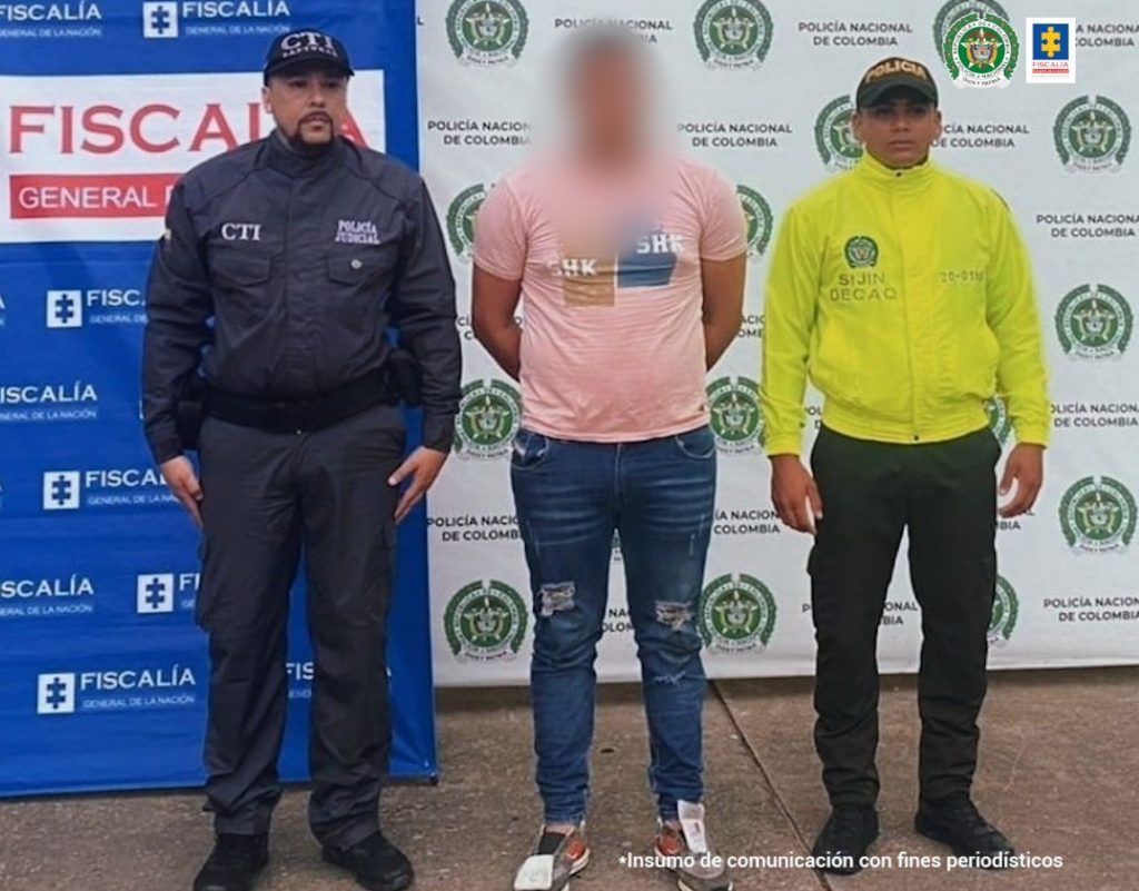 en la imagen se ve una persona detenida bajo custodia de un investigador del CTI y de un uniformado de la Policía Nacional. Detrás suyo un backing de la Fiscalía.