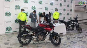 en la imagen se ven tres personas detenidas bajo custodia de dos integrantes de la Policía. Detrás suyo un backing institucional. Delante suyo varias motocicletas incautadas.
