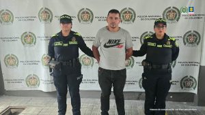 en la imagen se ve una persona detenida bajo custodia de dos uniformados de la Policía Nacional. Detrás suyo, un backing institucional.
