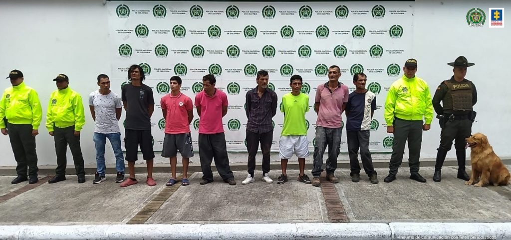 En la imagen se ven ocho personas detenidas en custodia de varios integrantes de la Policía Nacional. Detrás suyo un backing alusivo a la institución.