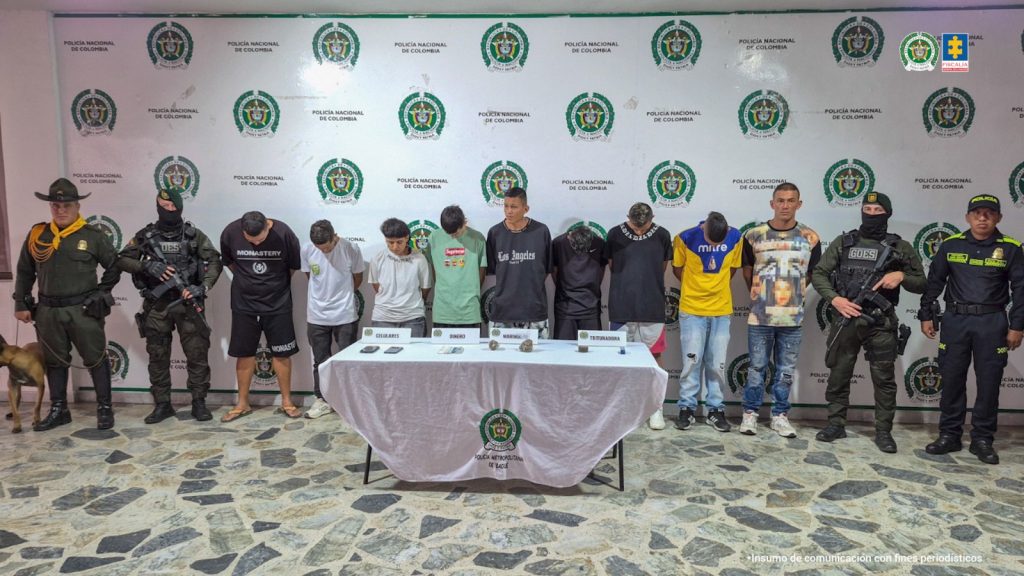 En la imagen se ve un grupo de personas detenidas bajo custodia de varios uniformados de la Policía Nacional. Delante de ellos una mesa con elementos incautados.