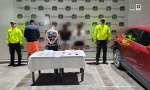 En la imagen se ven cuatro personas de espaldas, dos de ellas se ven pixeladas. Están bajo custodia de dos uniformados de la Policía Nacional. Al lado un carro decomisado y al frente una mesa con varios elementos incautados.