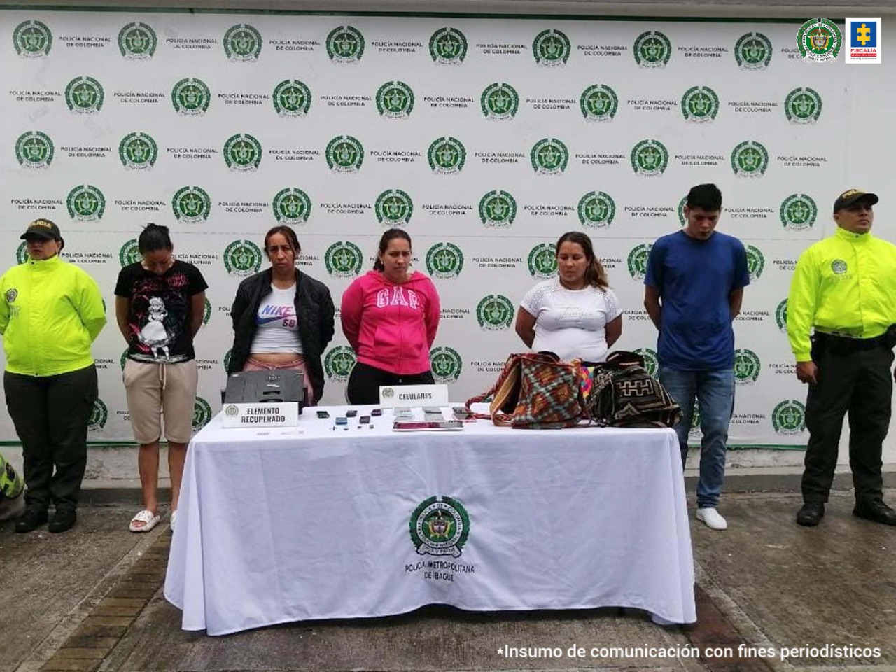 En la imagen se ven tres personas de la Policía Nacional de diferentes especialidades con un canino custodiando 5 personas detenidas. Adelante una mesa con varios elementos incautados como tabletas y morrales. Al fondo hay un backing de la Policía.