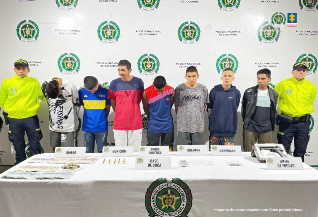 En la imagen se ven siete personas detenidas bajo custodia de integrantes de la Policía Nacional. Detrás suyo un backing institucional y delante de ellos una mesa con elementos incautados.